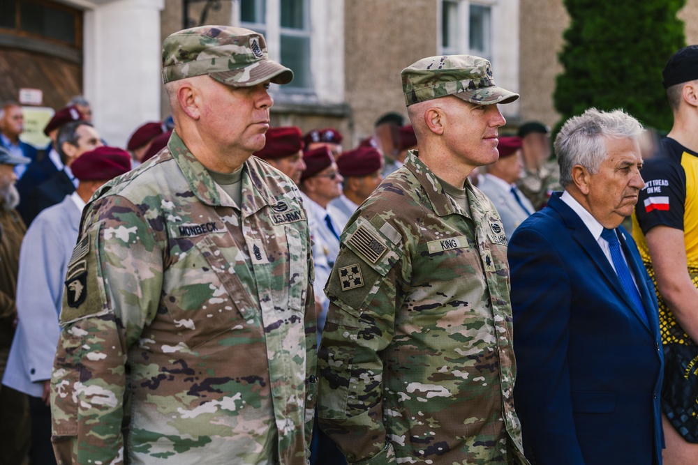 Two U.S. Army soldiers in MultiCam (OCP) camouflage uniforms stand at attention during what appears to be a formal military ceremony or commemoration event. Name tapes visible on their uniforms read 'MONBECK' and 'KING', both bearing U.S. Army insignia. Behind them, a crowd including soldiers wearing maroon berets and civilians is visible.