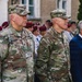 Two U.S. Army soldiers in MultiCam (OCP) camouflage uniforms stand at attention during what appears to be a formal military ceremony or commemoration event. Name tapes visible on their uniforms read 'MONBECK' and 'KING', both bearing U.S. Army insignia. Behind them, a crowd including soldiers wearing maroon berets and civilians is visible.