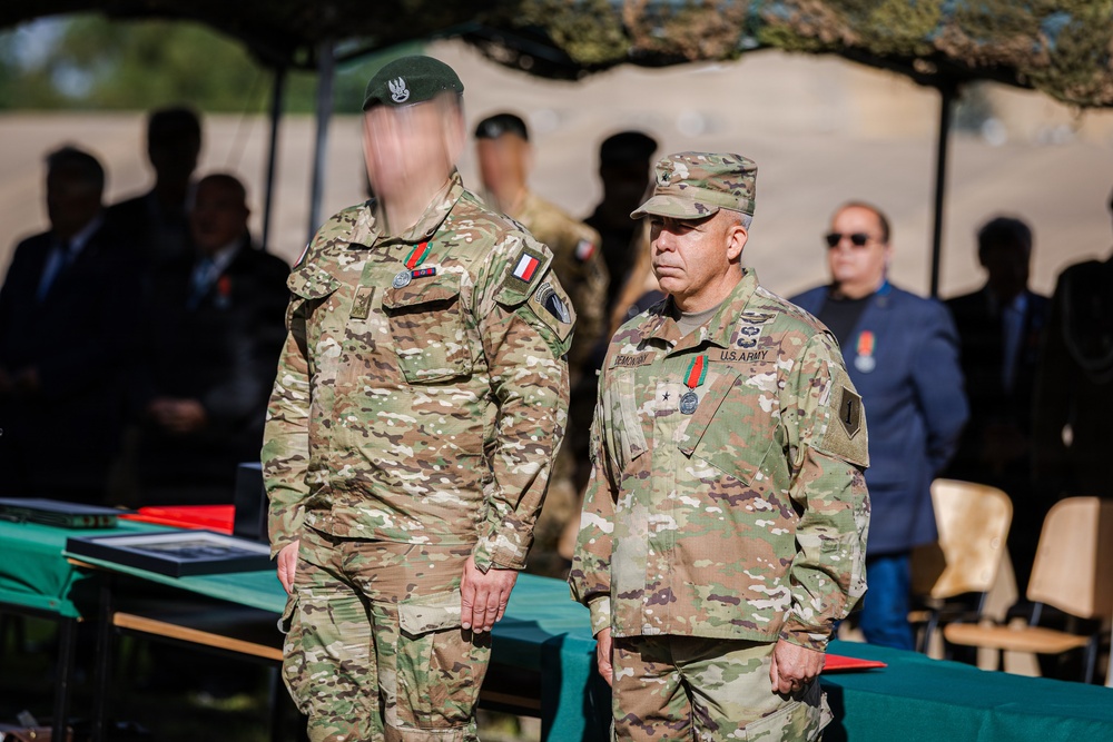Two military personnel in MultiCam/OCP camouflage uniforms are standing at what appears to be an outdoor military ceremony or award presentation. The soldier on the right wears a U.S. Army uniform with a general officer rank insignia (one star visible on cap) and a name tape reading 'DEMONAY' or similar. The soldier on the left wears a green beret and has their face digitally obscured, with what appears to be a French flag patch on the sleeve.