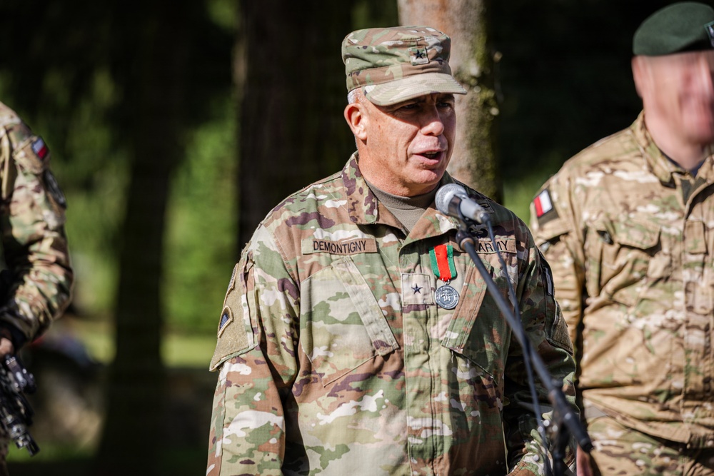 A U.S. Army Major General named Demontigny is speaking at a microphone during what appears to be an outdoor military ceremony or briefing. He is wearing OCP (Operational Camouflage Pattern) uniform with a two-star rank insignia on his cap and chest, along with a medal. Other military personnel in similar camouflage uniforms are visible in the background.