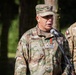 A U.S. Army Major General named Demontigny is speaking at a microphone during what appears to be an outdoor military ceremony or briefing. He is wearing OCP (Operational Camouflage Pattern) uniform with a two-star rank insignia on his cap and chest, along with a medal. Other military personnel in similar camouflage uniforms are visible in the background.