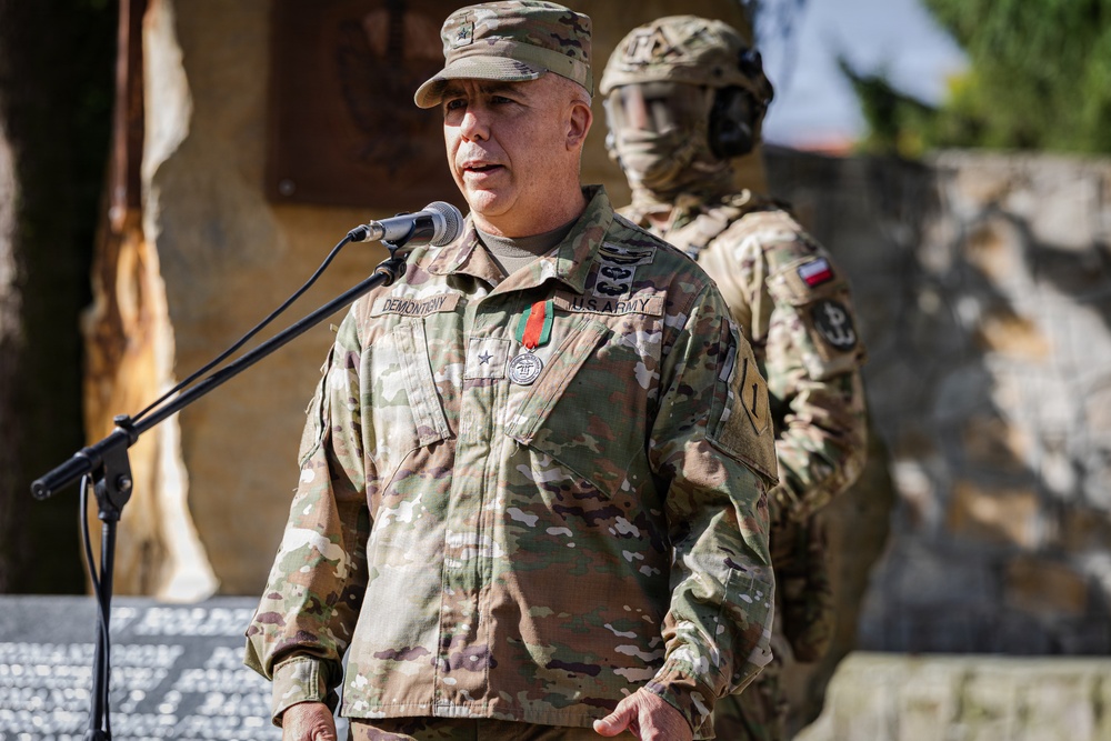 A U.S. Army Brigadier General with the name tape 'DEMONTEHY' is speaking at a microphone during what appears to be an official military ceremony. He is wearing OCP (Operational Camouflage Pattern) uniform with a single star rank insignia on his cap and a medal on his chest. A second soldier in full tactical gear, including a helmet and face covering, stands in the background.