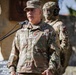 A U.S. Army Brigadier General with the name tape 'DEMONTEHY' is speaking at a microphone during what appears to be an official military ceremony. He is wearing OCP (Operational Camouflage Pattern) uniform with a single star rank insignia on his cap and a medal on his chest. A second soldier in full tactical gear, including a helmet and face covering, stands in the background.
