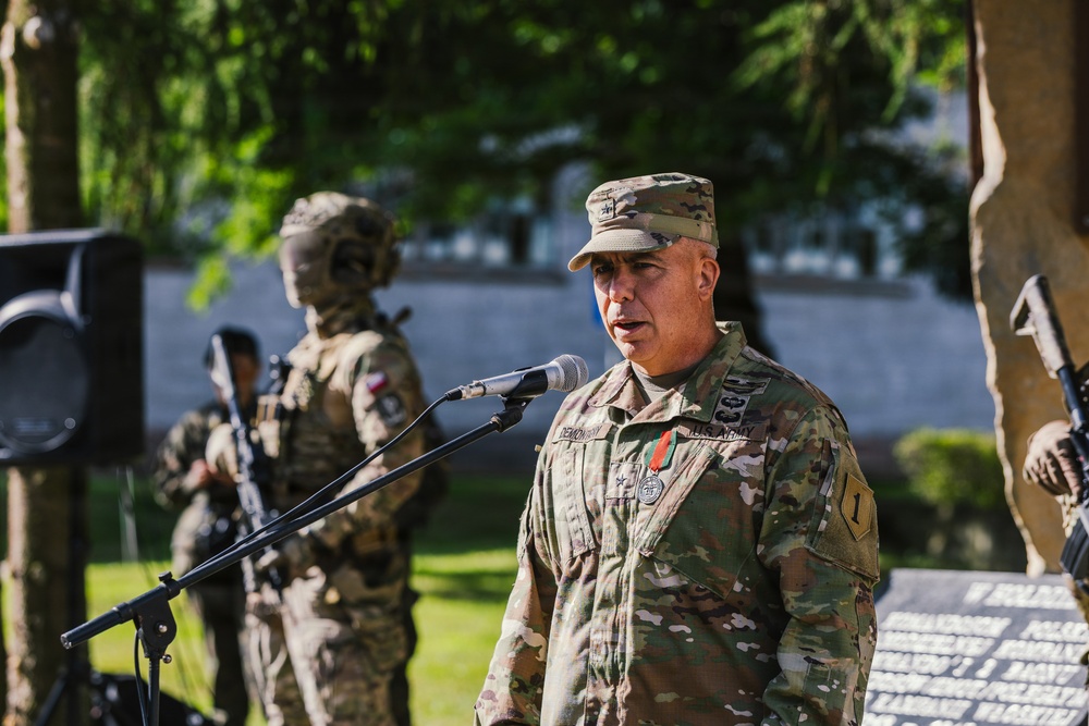 A U.S. Army officer wearing the rank of Brigadier General (one star) is speaking at a microphone during what appears to be an outdoor military ceremony. He is wearing MultiCam/OCP camouflage uniform with a medal and a '1' shoulder patch (1st Infantry Division). In the background, soldiers in tactical gear are visible, along with what appears to be a memorial monument with text in Polish.