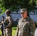 A U.S. Army officer wearing the rank of Brigadier General (one star) is speaking at a microphone during what appears to be an outdoor military ceremony. He is wearing MultiCam/OCP camouflage uniform with a medal and a '1' shoulder patch (1st Infantry Division). In the background, soldiers in tactical gear are visible, along with what appears to be a memorial monument with text in Polish.
