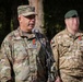 Two military personnel in camouflage uniforms are standing outdoors, with the foreground figure speaking at a microphone. The primary subject wears a US Army uniform with the name tape 'DEMONTIGNY' and a one-star general rank insignia on his cap. Both individuals are wearing matching medals with red and green ribbons.