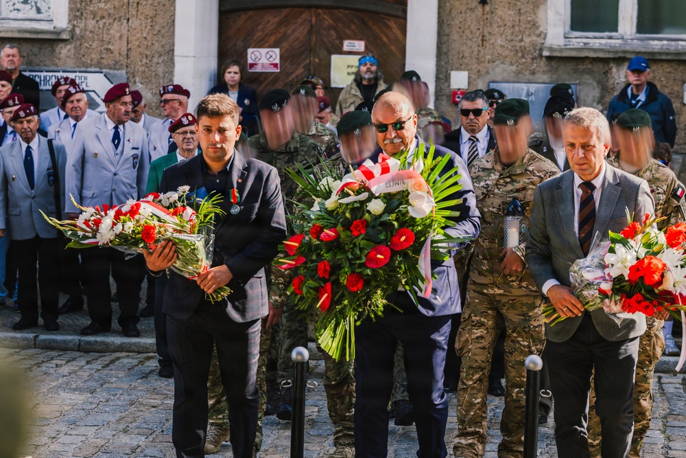 A formal commemorative ceremony is taking place, likely in Poland, where civilians and military personnel are carrying floral wreaths in red and white colors. Veterans wearing white jackets and maroon berets stand in formation in the background, alongside soldiers in camouflage uniforms. The setting appears to be in front of a historic stone building on a cobblestone square.
