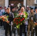 A formal commemorative ceremony is taking place, likely in Poland, where civilians and military personnel are carrying floral wreaths in red and white colors. Veterans wearing white jackets and maroon berets stand in formation in the background, alongside soldiers in camouflage uniforms. The setting appears to be in front of a historic stone building on a cobblestone square.