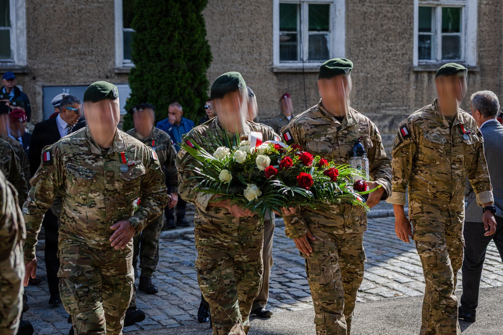 A group of military personnel in multicam camouflage uniforms and green berets are participating in what appears to be a wreath-laying ceremony. Two soldiers are carrying a large floral arrangement of red and white roses, suggesting a commemorative or memorial event. Civilians and other military personnel are visible in the background near a stone building.