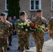 A group of military personnel in multicam camouflage uniforms and green berets are participating in what appears to be a wreath-laying ceremony. Two soldiers are carrying a large floral arrangement of red and white roses, suggesting a commemorative or memorial event. Civilians and other military personnel are visible in the background near a stone building.