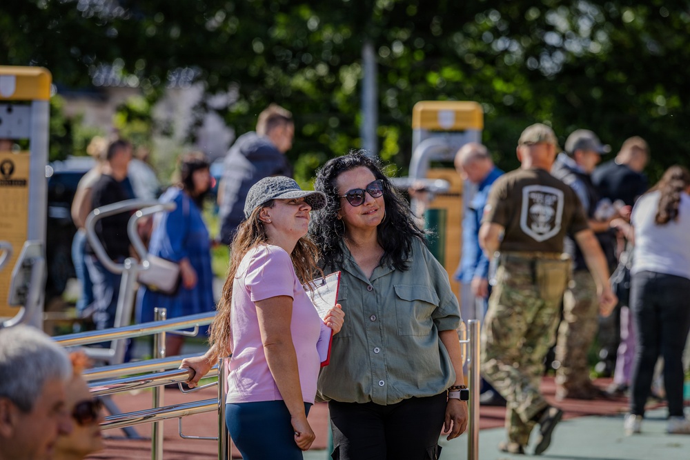 Two women stand together in the foreground of what appears to be an outdoor public fitness area or park. One woman wears a pink t-shirt and bucket hat, while the other wears a green shirt and sunglasses. In the background, a crowd of people mingles near outdoor exercise equipment, including one individual in military-style camouflage clothing.