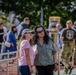 Two women stand together in the foreground of what appears to be an outdoor public fitness area or park. One woman wears a pink t-shirt and bucket hat, while the other wears a green shirt and sunglasses. In the background, a crowd of people mingles near outdoor exercise equipment, including one individual in military-style camouflage clothing.