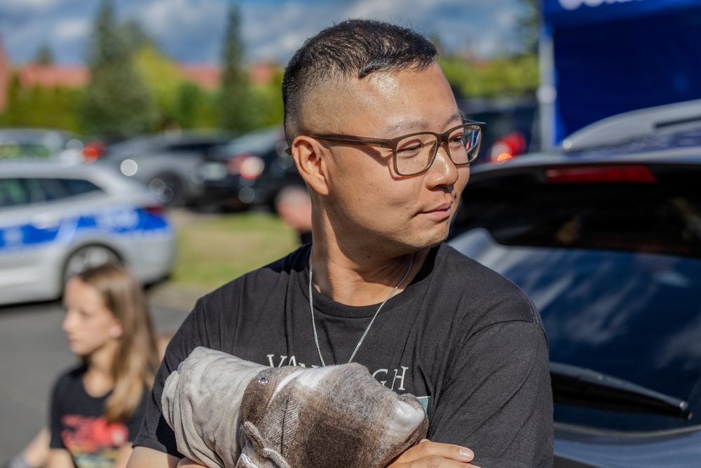 A man wearing glasses and a dark t-shirt stands outdoors in what appears to be a parking lot event, holding a gray cloth or garment. He is looking to his right with a neutral expression. The background shows parked cars, a blue tent or canopy, trees, and a child standing nearby.