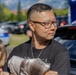A man wearing glasses and a dark t-shirt stands outdoors in what appears to be a parking lot event, holding a gray cloth or garment. He is looking to his right with a neutral expression. The background shows parked cars, a blue tent or canopy, trees, and a child standing nearby.