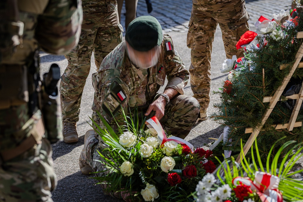 A soldier in multicam/camouflage uniform wearing a green beret is kneeling to lay a floral wreath during what appears to be a military commemoration ceremony. The soldier's uniform displays patches consistent with the Polish flag (white and red). Several other uniformed personnel stand nearby, and multiple wreaths with red and white ribbons are visible.