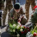 A soldier in multicam/camouflage uniform wearing a green beret is kneeling to lay a floral wreath during what appears to be a military commemoration ceremony. The soldier's uniform displays patches consistent with the Polish flag (white and red). Several other uniformed personnel stand nearby, and multiple wreaths with red and white ribbons are visible.