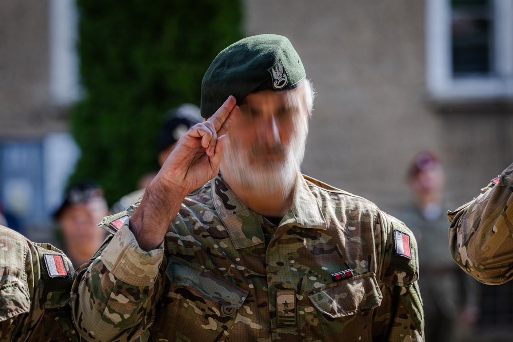 A military soldier in camouflage uniform is captured mid-salute, with motion blur obscuring the face. The soldier wears a green beret with a badge and displays Polish flag patches on the uniform sleeve. The setting appears to be an outdoor military ceremony or formal event.