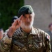 A military soldier in camouflage uniform is captured mid-salute, with motion blur obscuring the face. The soldier wears a green beret with a badge and displays Polish flag patches on the uniform sleeve. The setting appears to be an outdoor military ceremony or formal event.