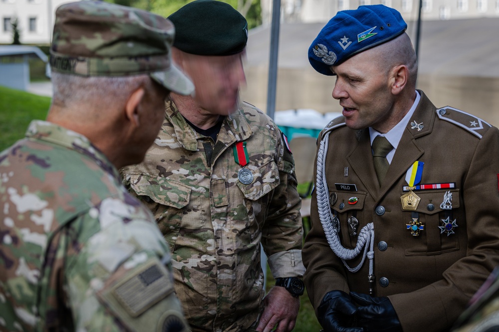 Three military personnel from different nations are engaged in conversation at what appears to be an outdoor military event or ceremony. The individuals wear distinct uniforms representing at least two different armed forces, with one in US Army OCP camouflage and another in a formal dress uniform with a blue beret and multiple decorations. The setting appears to be a military installation or ceremonial grounds.