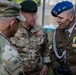 Three military personnel from different nations are engaged in conversation at what appears to be an outdoor military event or ceremony. The individuals wear distinct uniforms representing at least two different armed forces, with one in US Army OCP camouflage and another in a formal dress uniform with a blue beret and multiple decorations. The setting appears to be a military installation or ceremonial grounds.