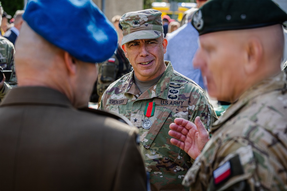 A U.S. Army officer with the name tape 'DEMONTENY' and rank insignia of a Brigadier General (one star) is engaged in conversation with two other military personnel. He is wearing OCP (Operational Camouflage Pattern) uniform with a medal on his chest and U.S. Army tape. The other individuals appear to be from allied nations, one wearing a blue beret and another wearing a dark green beret with a Polish flag patch visible.