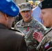 A U.S. Army officer with the name tape 'DEMONTENY' and rank insignia of a Brigadier General (one star) is engaged in conversation with two other military personnel. He is wearing OCP (Operational Camouflage Pattern) uniform with a medal on his chest and U.S. Army tape. The other individuals appear to be from allied nations, one wearing a blue beret and another wearing a dark green beret with a Polish flag patch visible.