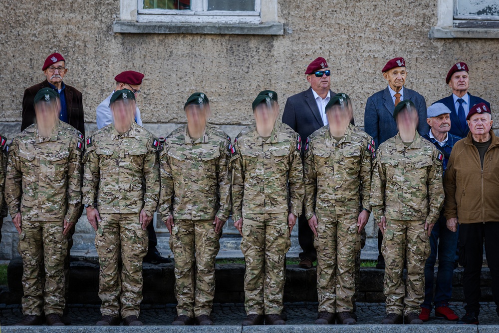 A formal military formation featuring soldiers in multicam camouflage uniforms with green berets, standing at attention in front of a stone building. Behind them stand several older civilians and veterans wearing maroon/burgundy berets, suggesting a ceremonial or commemorative event. The faces of the active-duty soldiers have been digitally obscured for privacy.