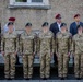 A formal military formation featuring soldiers in multicam camouflage uniforms with green berets, standing at attention in front of a stone building. Behind them stand several older civilians and veterans wearing maroon/burgundy berets, suggesting a ceremonial or commemorative event. The faces of the active-duty soldiers have been digitally obscured for privacy.