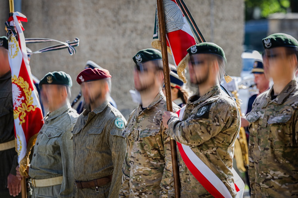 A military color guard ceremony featuring soldiers in various uniforms carrying flags with Polish national colors (red and white). The personnel wear different beret colors including green and maroon, along with camouflage and olive-drab uniforms bearing unit patches and eagle insignia. The setting appears to be an outdoor formal military parade or commemorative event.