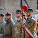 A military color guard ceremony featuring soldiers in various uniforms carrying flags with Polish national colors (red and white). The personnel wear different beret colors including green and maroon, along with camouflage and olive-drab uniforms bearing unit patches and eagle insignia. The setting appears to be an outdoor formal military parade or commemorative event.
