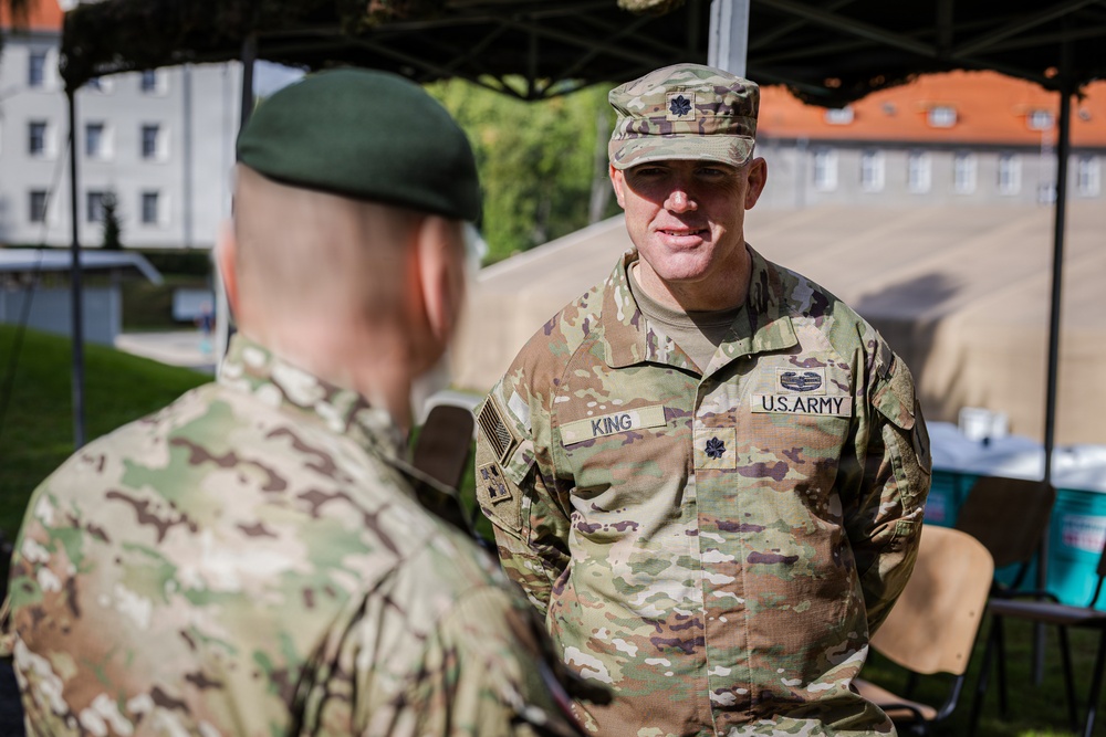 Two military personnel are engaged in conversation outdoors, likely at a military installation or exercise area. The soldier facing the camera wears U.S. Army OCP (Operational Camouflage Pattern) uniform with the name tape 'KING' and a Lieutenant Colonel oak leaf insignia. The other individual, seen from behind, wears a green beret suggesting possible special forces or allied military affiliation.