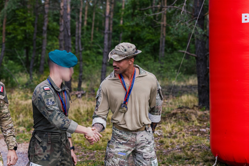 Two military personnel are shaking hands in an outdoor forested setting, both wearing medals on red and blue ribbons, suggesting a competition or ceremony. One individual wears Polish military camouflage with a teal beret and a Polish flag patch, while the other wears a MultiCam uniform with a patrol cap. A red inflatable structure is visible on the right side of the frame.
