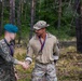 Two military personnel are shaking hands in an outdoor forested setting, both wearing medals on red and blue ribbons, suggesting a competition or ceremony. One individual wears Polish military camouflage with a teal beret and a Polish flag patch, while the other wears a MultiCam uniform with a patrol cap. A red inflatable structure is visible on the right side of the frame.