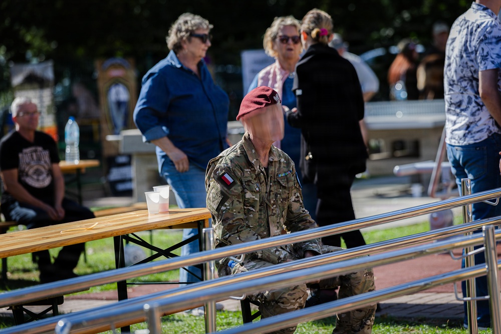 An outdoor public gathering or event featuring a soldier in camouflage uniform wearing a red/maroon beret, seated near metal railings and wooden picnic tables. The soldier has a flag patch visible on the sleeve, consistent with Polish military insignia. Several civilians are present in the background, suggesting a public commemoration or military-themed event.