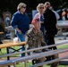 An outdoor public gathering or event featuring a soldier in camouflage uniform wearing a red/maroon beret, seated near metal railings and wooden picnic tables. The soldier has a flag patch visible on the sleeve, consistent with Polish military insignia. Several civilians are present in the background, suggesting a public commemoration or military-themed event.