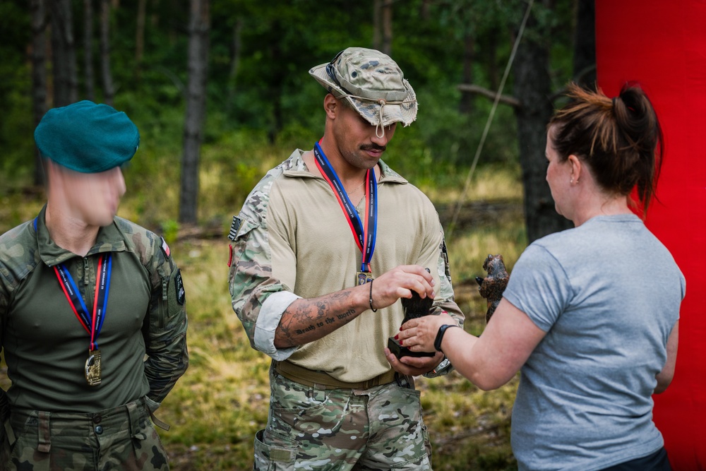 Three individuals are gathered outdoors in a forested setting, appearing to be at a military competition or awards ceremony. Two individuals in military uniforms are wearing medals on red-and-blue lanyards, while a civilian woman in a grey shirt appears to be presenting or exchanging a trophy or award. The setting suggests a post-competition recognition event.