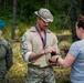 Three individuals are gathered outdoors in a forested setting, appearing to be at a military competition or awards ceremony. Two individuals in military uniforms are wearing medals on red-and-blue lanyards, while a civilian woman in a grey shirt appears to be presenting or exchanging a trophy or award. The setting suggests a post-competition recognition event.