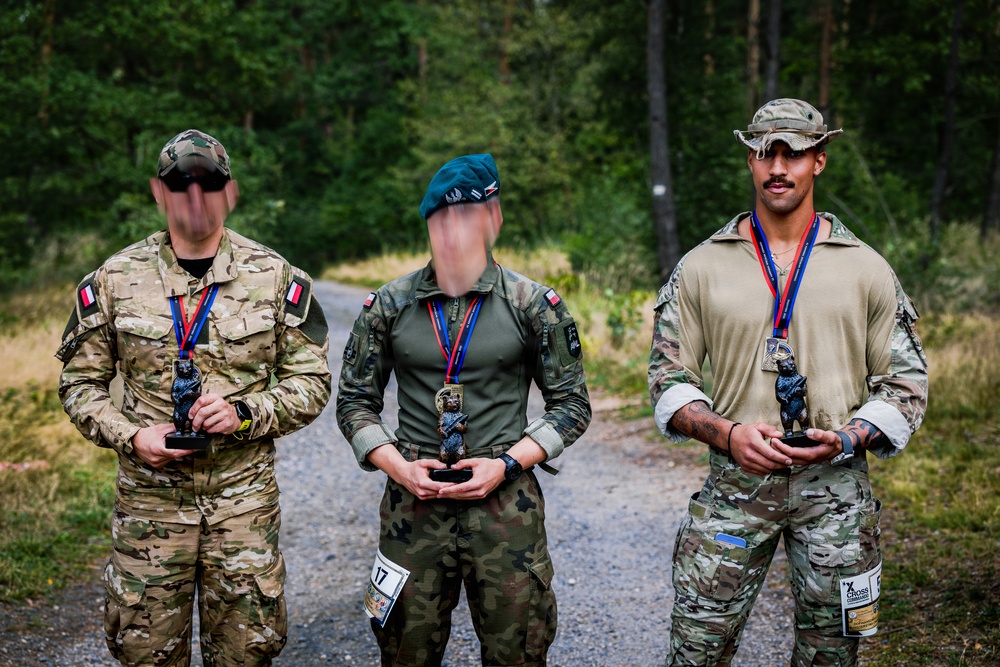 Three military personnel in different uniforms stand together on a gravel path in a forested area, each holding a trophy figurine and wearing a medal on a red-white-blue ribbon. The individuals appear to be posing for a post-competition photograph, likely following a military athletic or skills competition. Their faces have been digitally obscured for privacy.