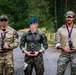 Three military personnel in different uniforms stand together on a gravel path in a forested area, each holding a trophy figurine and wearing a medal on a red-white-blue ribbon. The individuals appear to be posing for a post-competition photograph, likely following a military athletic or skills competition. Their faces have been digitally obscured for privacy.