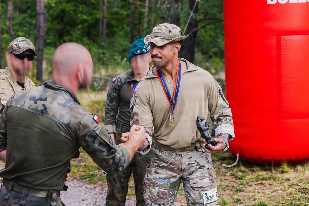 A military ceremony or competition award presentation is taking place outdoors near a forested area. A soldier in tan/multicam uniform wearing a medal and holding a small trophy figurine is shaking hands with another soldier in Polish camouflage pattern uniform. The event appears to be a multinational military competition based on visible insignia and the 'Cross Command' label on the recipient's leg.