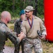 A military ceremony or competition award presentation is taking place outdoors near a forested area. A soldier in tan/multicam uniform wearing a medal and holding a small trophy figurine is shaking hands with another soldier in Polish camouflage pattern uniform. The event appears to be a multinational military competition based on visible insignia and the 'Cross Command' label on the recipient's leg.