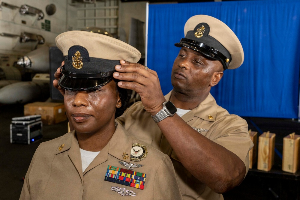 A U.S. Navy chief petty officer is having their combination cover (cap) placed on their head by another senior enlisted sailor during what appears to be a chief petty officer pinning or advancement ceremony. Both individuals are wearing the tan/khaki Service Uniform with chief petty officer insignia. The ceremony appears to be taking place in a ship's hangar bay, indicated by aircraft and equipment visible in the background.