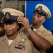 A U.S. Navy chief petty officer is having their combination cover (cap) placed on their head by another senior enlisted sailor during what appears to be a chief petty officer pinning or advancement ceremony. Both individuals are wearing the tan/khaki Service Uniform with chief petty officer insignia. The ceremony appears to be taking place in a ship's hangar bay, indicated by aircraft and equipment visible in the background.