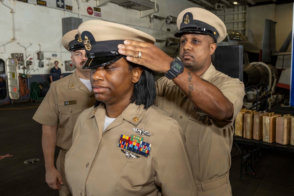 A U.S. Navy promotion or pinning ceremony is taking place in what appears to be a ship's hangar bay. A male sailor is placing a chief petty officer combination cover (hat) onto the head of a female sailor, who is visibly emotional. A third sailor stands in the background observing the ceremony.