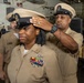 A U.S. Navy promotion or pinning ceremony is taking place in what appears to be a ship's hangar bay. A male sailor is placing a chief petty officer combination cover (hat) onto the head of a female sailor, who is visibly emotional. A third sailor stands in the background observing the ceremony.