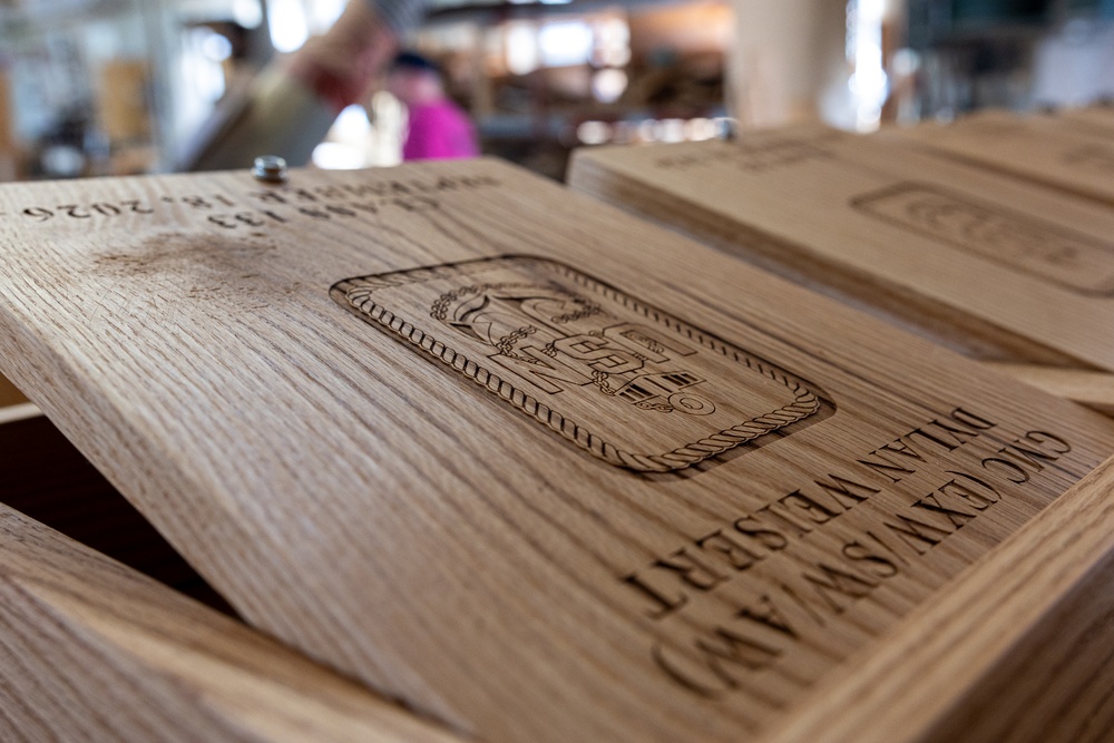 The image shows laser-engraved wooden boards or plaques stacked together, likely custom-made awards or commemorative items. The foreground board displays an engraved emblem/crest along with the text 'CIC/CXW/SW/AM' and 'DYLAN WEISERT', along with what appears to be a date '2026'. The setting appears to be a workshop or retail/display environment with blurred figures visible in the background.