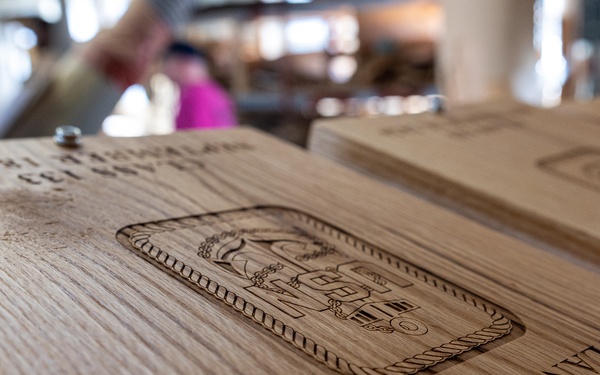 The image shows laser-engraved wooden boards or plaques stacked together, likely custom-made awards or commemorative items. The foreground board displays an engraved emblem/crest along with the text 'CIC/CXW/SW/AM' and 'DYLAN WEISERT', along with what appears to be a date '2026'. The setting appears to be a workshop or retail/display environment with blurred figures visible in the background.