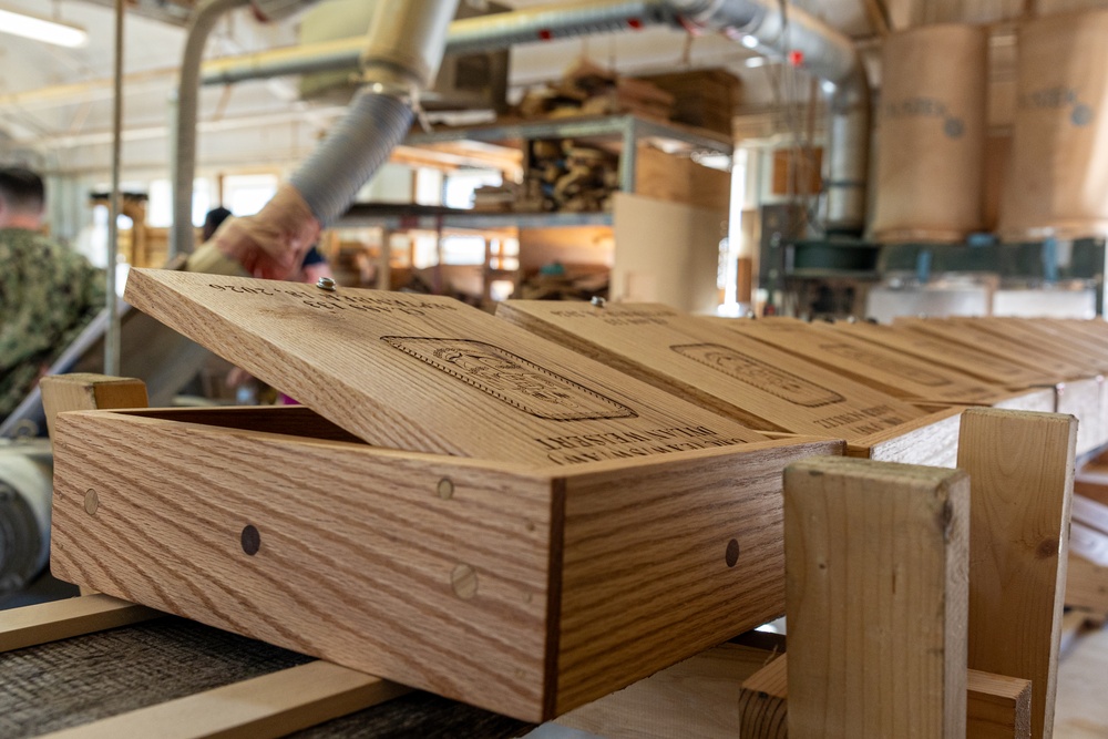 The image shows a woodworking workshop environment where laser-engraved wooden boxes are being produced on an assembly line or staging area. The foreground features a light oak wooden box with its lid open, displaying detailed engraved imagery and text on the lid surface. In the background, workers and workshop equipment are visible, along with additional completed boxes and raw materials.
