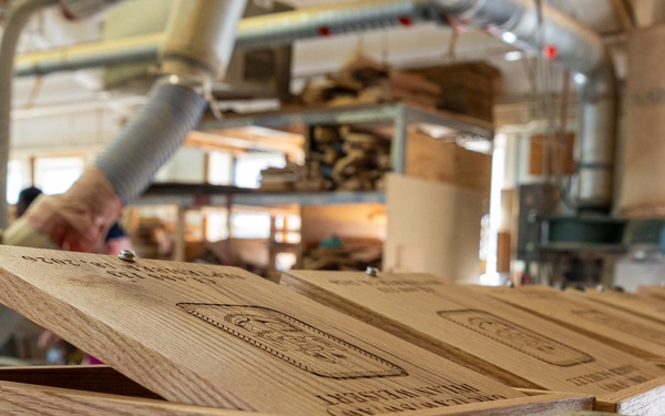 The image shows a woodworking workshop environment where laser-engraved wooden boxes are being produced on an assembly line or staging area. The foreground features a light oak wooden box with its lid open, displaying detailed engraved imagery and text on the lid surface. In the background, workers and workshop equipment are visible, along with additional completed boxes and raw materials.
