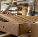 The image shows a woodworking workshop environment where laser-engraved wooden boxes are being produced on an assembly line or staging area. The foreground features a light oak wooden box with its lid open, displaying detailed engraved imagery and text on the lid surface. In the background, workers and workshop equipment are visible, along with additional completed boxes and raw materials.